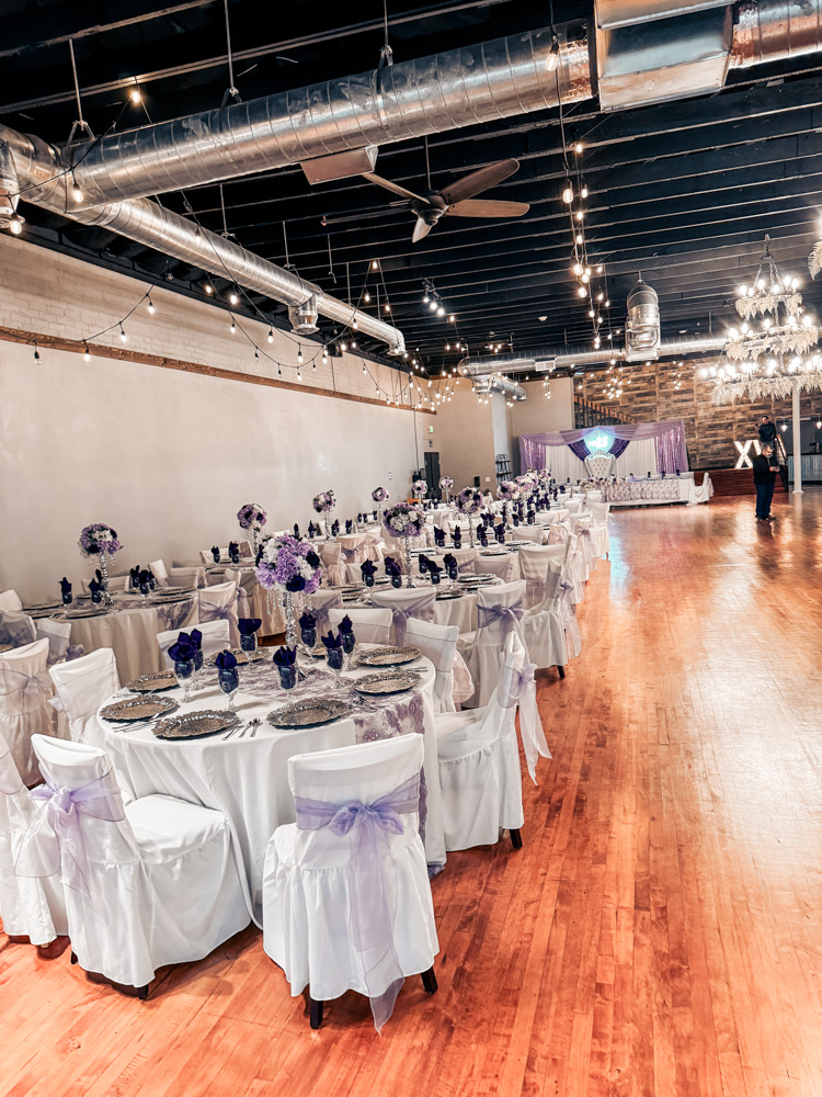 Romantic indoor wedding ceremony aisle featuring a white runner, candle-lit lanterns, and a beautifully draped floral altar.