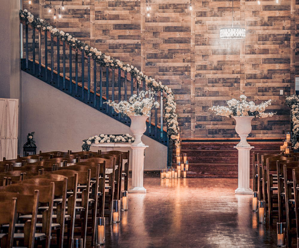 An indoor wedding ceremony aisle lined with glowing pillar candles in glass cylinders. In the background, a rustic wood-paneled wall stands behind a grand staircase draped in a thick white floral garland. Two large white classical urns on pedestals filled with white flowers frame the altar area.