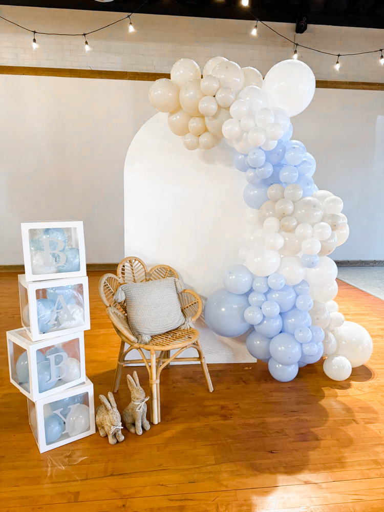 A baby shower photo backdrop featuring a large white arched panel adorned with a blue, white, and cream balloon garland. To the left, white stacked boxes spell "BABY" filled with matching balloons. A rattan chair with a grey pillow sits in front, flanked by two small bunny figurines on a light wood floor.