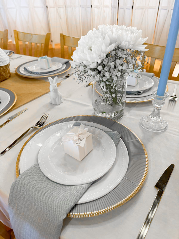 A refined table setting featuring a white tablecloth, grey and gold-rimmed chargers, and a white "Thank You" favor box. A crystal vase with white hydrangeas and baby's breath serves as a centerpiece alongside a light blue taper candle and a small white bunny figurine by venue at sombras