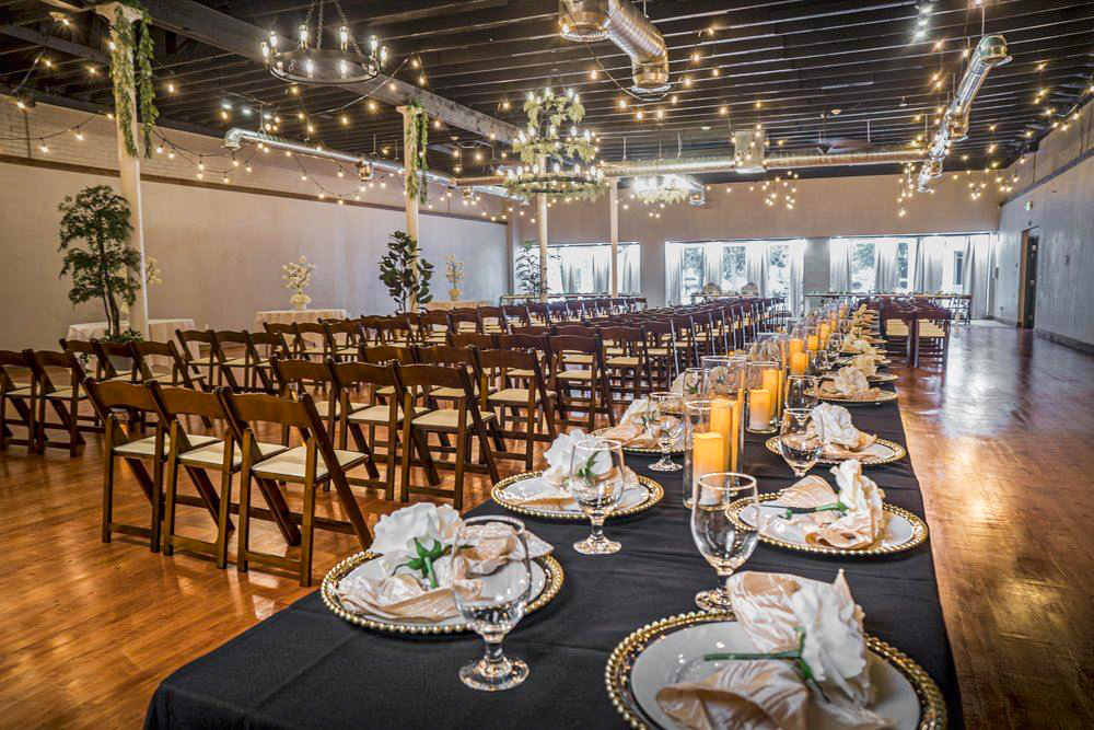 A wide view of an indoor venue event space with dark wood ceilings, exposed ductwork, and hanging Edison string lights. Rows of dark wood folding chairs face away from the camera, while a long banquet table in the foreground is set with black linens, gold-rimmed plates, white floral centerpieces, and tall amber candles.