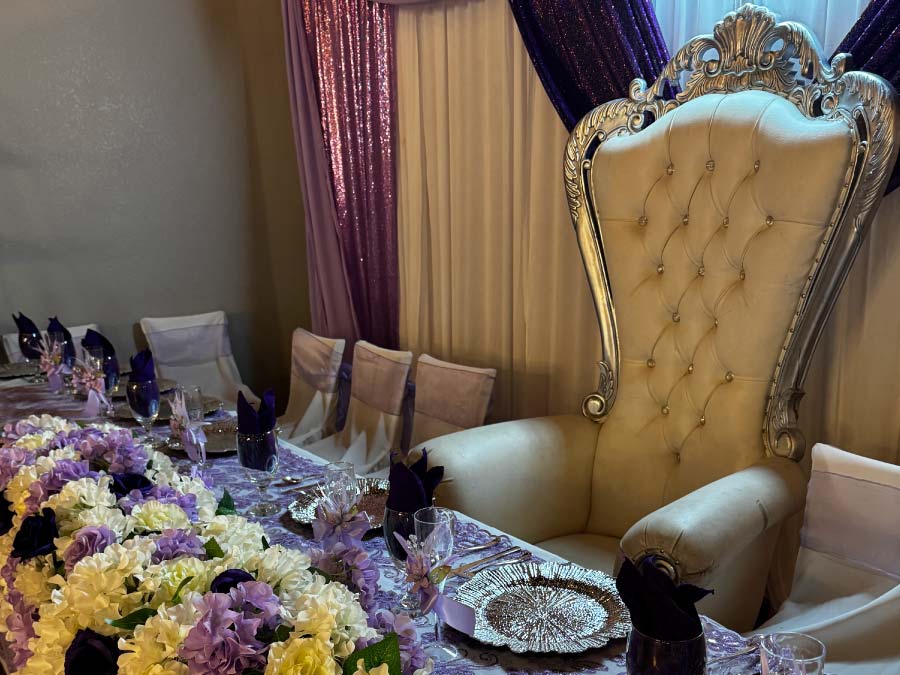 A luxurious Quinceañera head table featuring a large, cream-colored tufted throne with a silver ornate frame, set against a backdrop of white and sparkling lavender drapes. The table is adorned with a lush runner of white and purple flowers, elegant silver chargers, and deep purple napkins