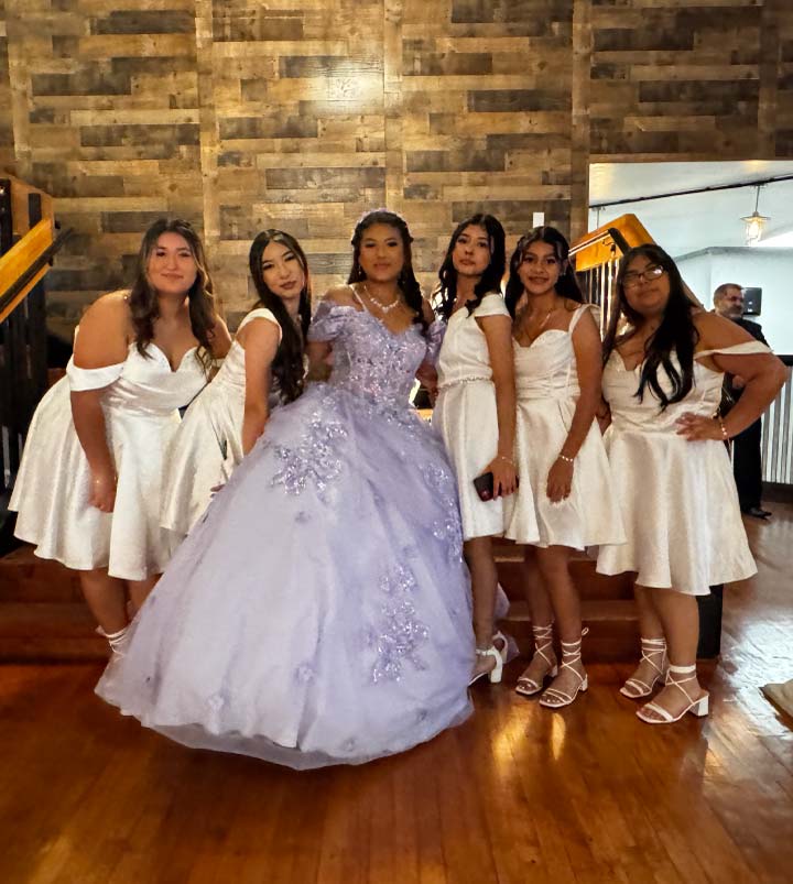A Quinceañera in a voluminous lavender ballgown embellished with floral appliqués stands smiling with her court of six young women in matching white off-the-shoulder cocktail dresses. They are posed in front of a rustic wood-paneled wall inside a modern quinceañera venue.