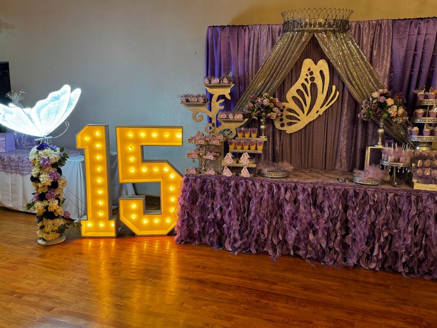A vibrant dessert and display table for a Quinceañera featuring a purple ruffled tablecloth and a lavender sequin backdrop with a large golden butterfly. The setup includes glowing "15" marquee numbers, tiered cupcake stands, and themed treats, all arranged on a polished hardwood floor.